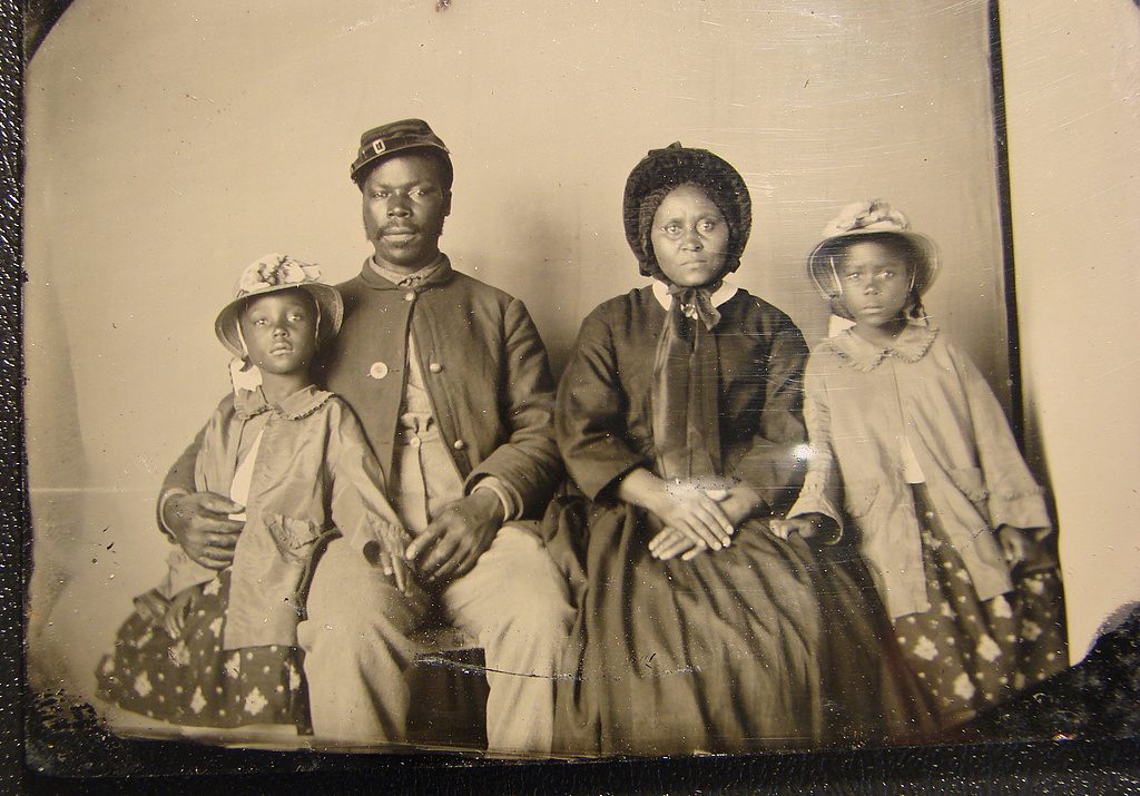 A black man in uniform and a black woman wearing a dress and bonnet sitting for a portrait with their two daughters on each side.