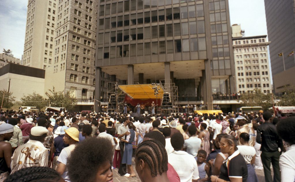A crowd of African American people looking towards a stage set up in front of a large modern office building.
