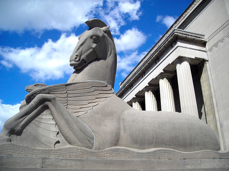 A stone sculpture of a horse sitting on an abstract wave with columns in the background.